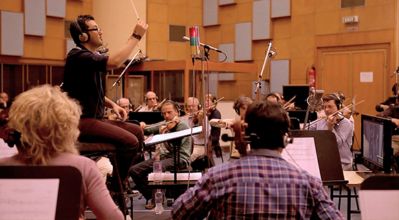 A conductor with headphones directs an orchestra in a wood-paneled recording studio.