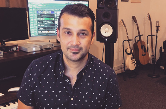 A man in a patterned shirt sits in a music studio with guitars and studio monitors behind him.