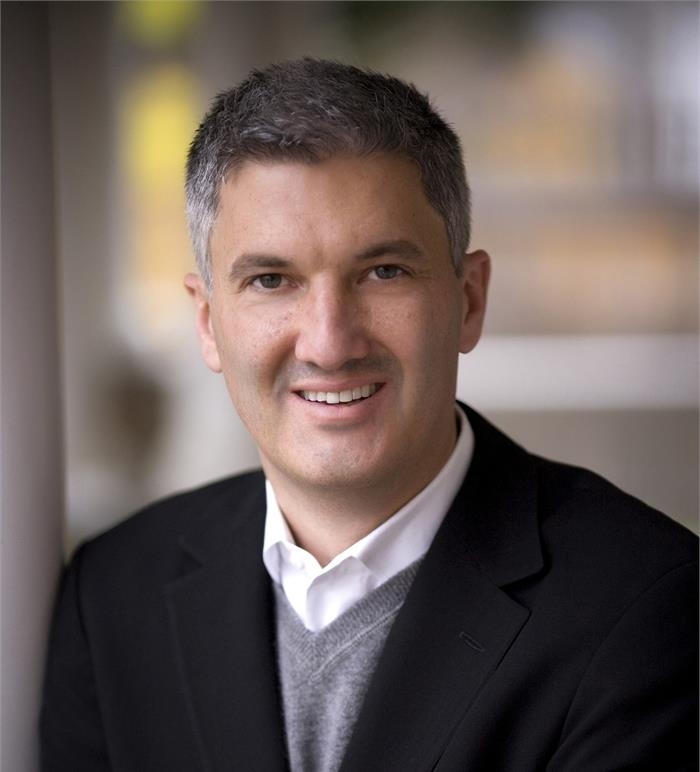 A smiling man with short salt-and-pepper hair wearing a black blazer over a white shirt and grey sweater.