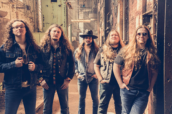 Five men with long hair posing in a weathered, industrial hallway.