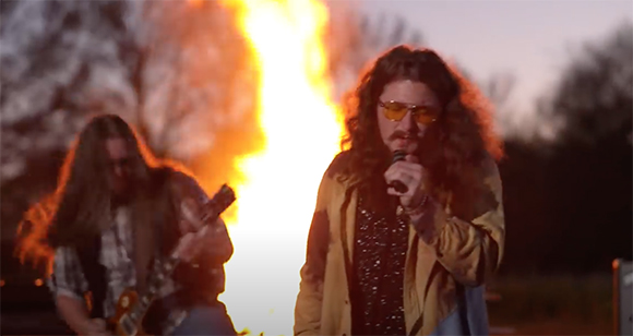 A man with long hair sings into a microphone outdoors at night with a large fire behind him.