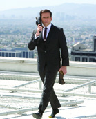 A man in a dark suit walks across a rooftop while talking on a mobile phone against a city skyline.
