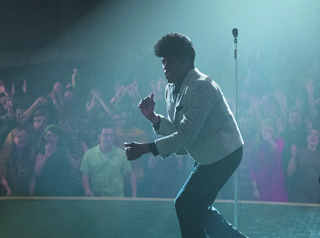 A man with an afro wearing a light patterned blazer dances on a stage before a crowd of people.