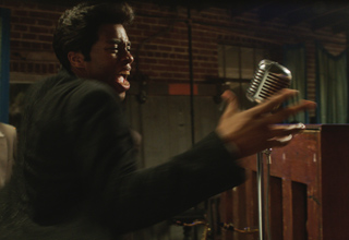 A man in a dark suit sings with an open mouth toward a silver vintage microphone.