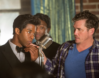 A man in a tuxedo looks skeptically at a man in a plaid shirt near a vintage microphone.
