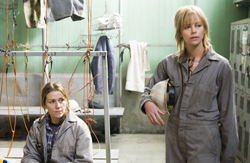 Two women in grey work clothes stand in a room with hanging gear and lockers.