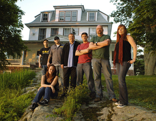 Six people pose together on a grassy lawn in front of a large, two-story house.
