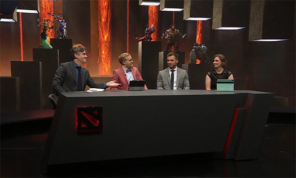 Four people in formal attire sit behind a desk in a studio decorated with video game character statues.