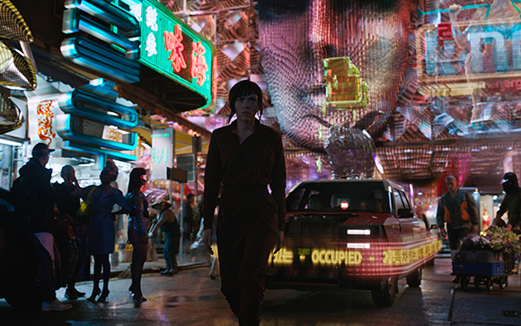 A woman walks through a crowded, neon-lit street with large digital displays and a passing car.