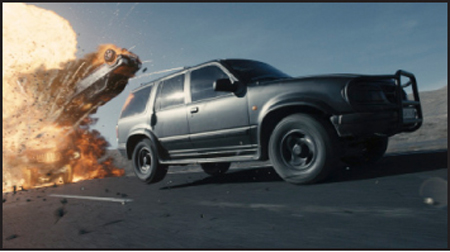 A black SUV speeds along a desert road as a massive fireball erupts behind it.