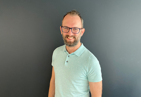 A smiling man with glasses and a beard wearing a light green polo shirt against a dark gray background.