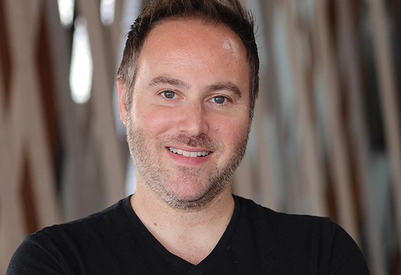 A smiling man with short brown hair and light stubble wearing a black v-neck shirt.