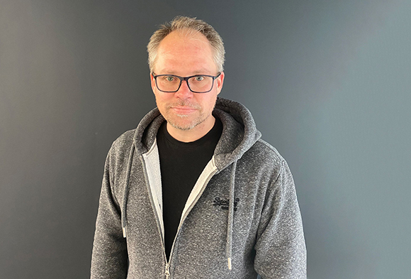 A man with short light hair and glasses wearing a grey zip-up hoodie over a black t-shirt.