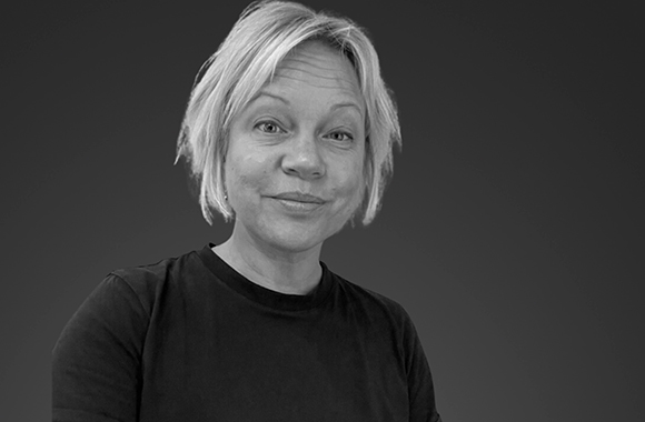 A black and white portrait of a woman with short blonde hair wearing a dark t-shirt against a dark background.