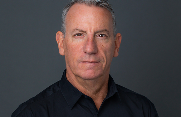 A middle-aged man with short graying hair and brown eyes wearing a black button-down shirt against a dark gray background.