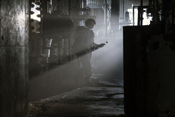 Silhouette of a person carrying a rifle through a dark, misty industrial facility with light beams.