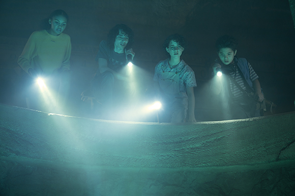Four children use flashlights to illuminate a dark, misty cave.