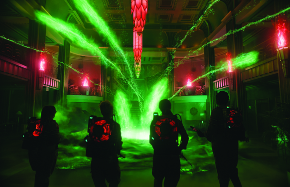 Four silhouetted figures with glowing red backpacks face a massive green energy vortex in a large hall.