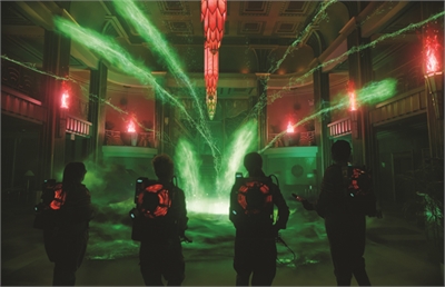 Four silhouetted figures in gear face a large explosion of green light and energy in a grand hall.