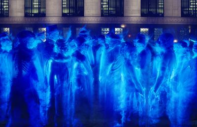 A crowd of people dressed in costumes, glowing in intense blue light against a dark building.