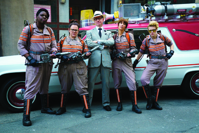 Five people in Ghostbusters jumpsuits and a man in a grey suit pose in front of a white car.