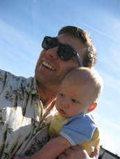 A smiling man wearing sunglasses holds a baby against a clear blue sky.