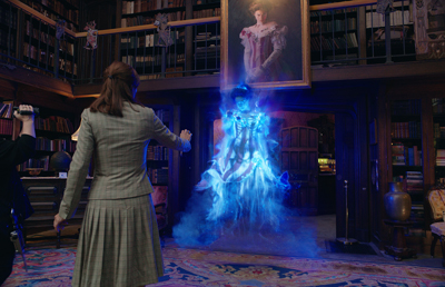 A woman in a grey skirt suit stands before a glowing, translucent blue spirit in a dark library.