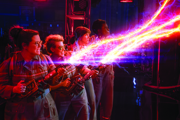 Four women aim glowing, energy-emitting proton packs toward a bright light in a dark room.