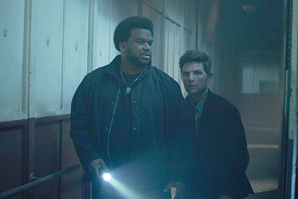 A man in a dark jacket holds a bright flashlight while walking through a dimly lit hallway with another man.