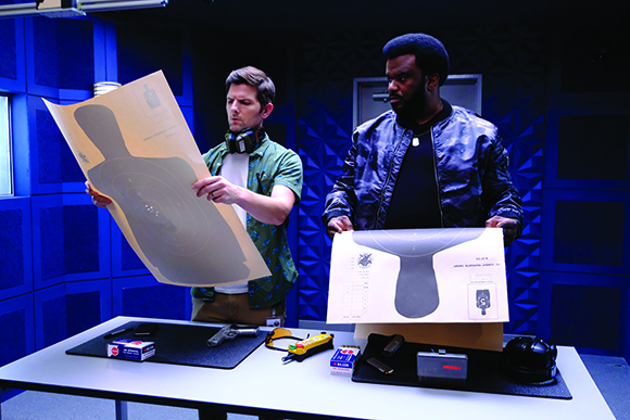 Two men stand behind a table covered in technical diagrams, tools, and equipment in a blue-lit room.