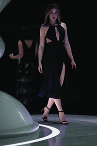 A woman in a long black halter dress with cutouts walks forward on a dark runway.