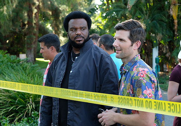 Two men stand behind yellow police line tape in a lush, outdoor setting.