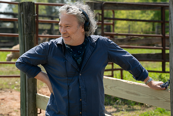 A man with gray hair and headphones wearing a blue button-down shirt stands outdoors by a fence.