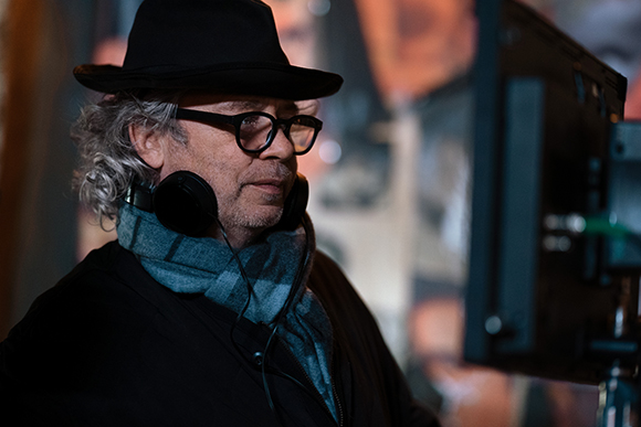 A man wearing a black fedora, glasses, and headphones with a blue scarf looks toward a monitor.