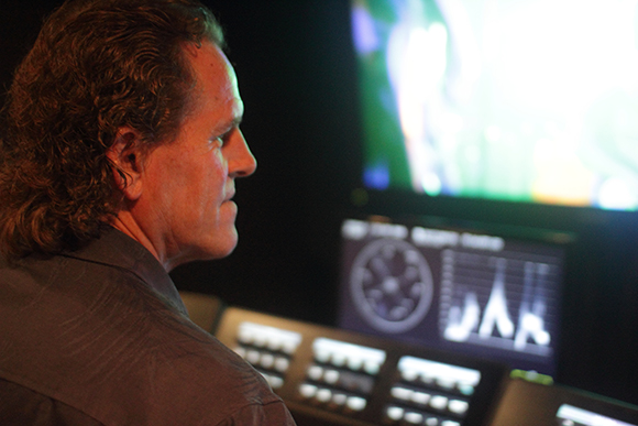 Side profile of a man watching a large glowing screen and monitoring data on a control panel in a dark room.