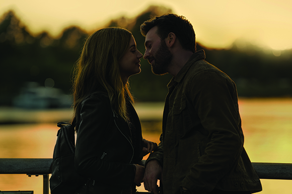 A man and a woman stand close together, foreheads nearly touching, against a sunset background.