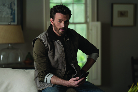 A man with dark hair and a beard sits indoors, wearing a brown vest and holding a smartphone.
