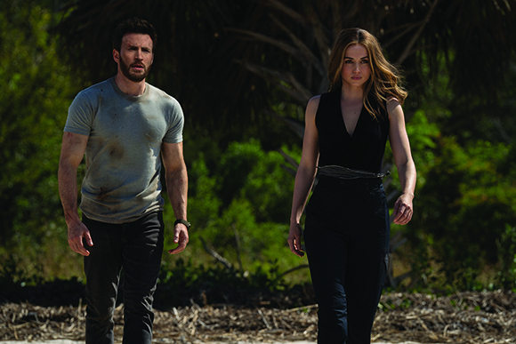 A man in a grey t-shirt and a woman in a black sleeveless top walk through a wooded area.