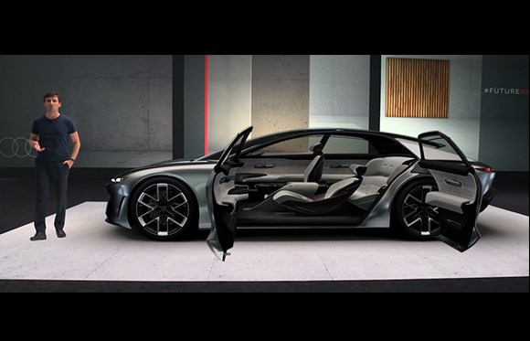 A man in a dark t-shirt stands beside a silver concept car with its doors open on a white platform.
