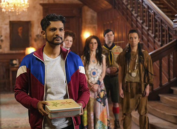 A man in a red and blue jacket holds a decorative box, standing before four people in a wood-paneled room.