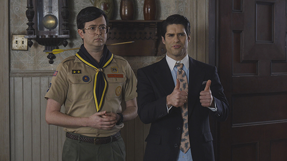 A man in a tan scout uniform stands next to a man in a dark suit giving two thumbs up.