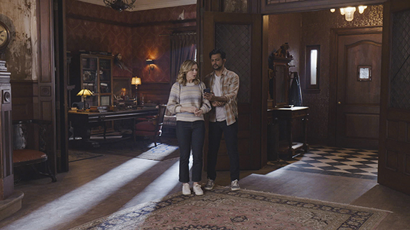 A man and woman stand in a vintage-style room with patterned wallpaper and dark wood furniture.