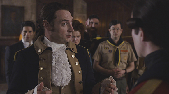 A man in a 18th-century style tan and navy coat looks intensely at someone off-camera.