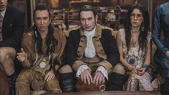 Four people in historical costumes sit together on a couch indoors.