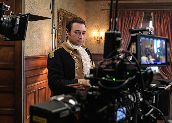 An actor in a dark blue military uniform stands behind a professional film camera on a set.