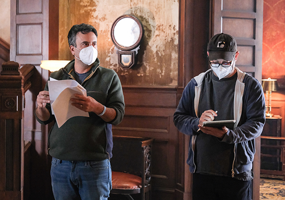 Two men wearing white face masks stand in a room; one holds papers and the other writes in a notebook.