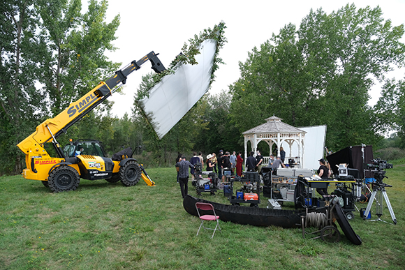 A yellow telehandler lifts a large white sheet over a film crew and equipment in a grassy field.