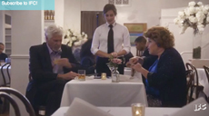 A server in a white shirt and black tie stands between two seated diners at a restaurant table.
