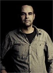 A man in a light-colored button-down shirt poses against a dark background.