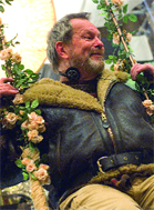 A bearded man in a dark shearling jacket sits behind climbing rose bushes.